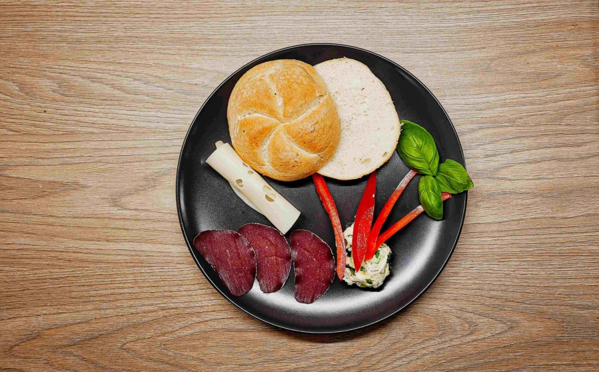 Na czarnym talerzu leży przekrojona bułka, ser żółty, salami, masło czosnkowe, papryka w paskach i liście bazylii